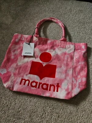 Isabel Marant Sac Yenky Tie Dye - Trendig shoppingväska i rosa och vit tie-dye med stort rött Isabel Marant-tryck på framsidan. Väskan har två rejäla handtag och är tillverkad i slitstark canvas. Perfekt för att bära allt du behöver till skolan, stranden eller stan.