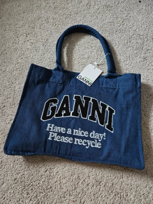Blå tote bag från Ganni - Snygg blå shoppingväska från Ganni i kraftigt tyg med broderad text 'GANNI Have a nice day! Please recycle' på framsidan. Väskan har två rejäla handtag och en stilren, rektangulär form. Perfekt för att bära allt du behöver till skolan eller stan.