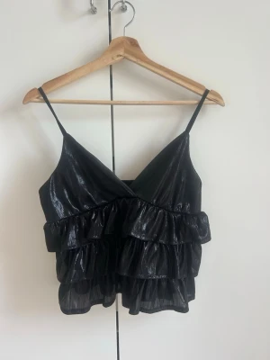 Svart glittrig volangtop med smala band - Snygg svart topp med glittrigt tyg och flera volanglager. Toppen har smala axelband, omlott framtill och en croppad passform som ger en trendig look. Perfekt för dig som vill sticka ut lite extra.