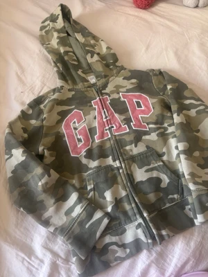 Camo hoodie med GAP-tryck - Säljer en camouflagemönstrad hoodie från GAP med dragkedja och stor huva. Framsidan har ett stort rosa GAP-tryck i college-stil. Hoodien har kängurufickor och är perfekt för en avslappnad streetwear-look. Köpt på Vinted står kids M 