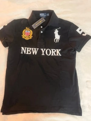 Svart pikétröja Polo Ralph Lauren - Snygg svart pikétröja från Polo Ralph Lauren med vit broderad logga, texten NEW YORK framtill och gul broderad detalj på bröstet. Kortärmad modell med krage och knappar. Perfekt för en sportig och stilren look.