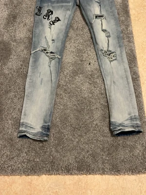 amiri Ljusblå slitna jeans med tryck - Säljer ett par amiri ljusblå jeans med slitningar och hål på båda benen. Jeansen har svarta tryckdetaljer på vänster lår och är i en rak, smal passform. Perfekta för dig som gillar en edgy och trendig stil.