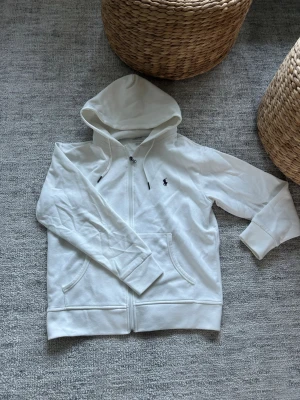 Vit hoodie med dragkedja och huva - Säljer en stilren vit hoodie med dragkedja och huva. Hoodien har två fickor framtill och en liten broderad logga på bröstet. Tillverkad i mjukt material som känns skönt mot huden. Perfekt för en avslappnad look.