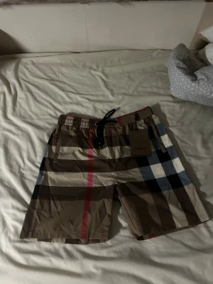 Burberry shorts - Snygga badbyxor från Burberry med klassiskt beige, svart, vitt och rött rutmönster. De har elastisk midja med svart dragsko och en bakficka. Perfekta för stranden eller poolen och ger en lyxig känsla till din sommarstil.