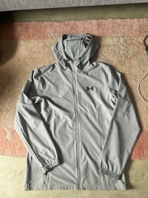 Under Armour jacka - Under Armour jacka med huva och dragkedja framtill. Jackan har resår i ärmslut med texten "UNDER ARMOUR" och en diskret logotyp på bröstet. Kvitto finns kvar digitalt på mail, pris kan diskuteras!! Tveka inte att skriva!!