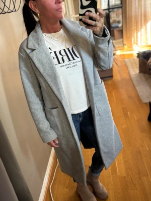 Ljusgrå lång kappa med fickor - Snygg ljusgrå kappa i rak modell med stora utanpåfickor och bred krage. Kappan är knälång och har en stilren design som passar perfekt till jeans och boots. Materialet ser ut att vara ullblandning för en mjuk och varm känsla. Storlek XS men passar mig som har st:S