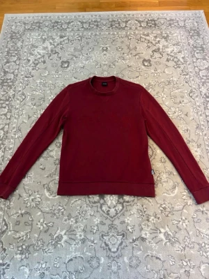 Mörkröd sweatshirt från Hugo boss - En stilren mörkröd sweatshirt med rund halsringning och långa ärmar. Tröjan har ribbade muddar vid ärmslut och nederkant. Perfekt för lager-på-lager och enkel att matcha med olika outfits.