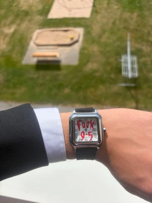 ”Fuck 9-5” klocka - Säljer en stilren klocka med svart armband och fyrkantigt silverfärgat boett. Urtavlan har romerska siffror och ett unikt tryck med texten 'Fuck 9-5' i rött. Perfekt för dig som vill sticka ut och visa att du går din egen väg.