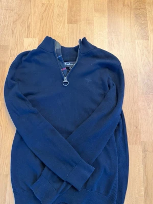 Mörkblå halvzip-tröja från Barbour - Snygg mörkblå stickad tröja från Barbour med halv dragkedja och ribbade muddar. Klassisk design med diskret logga på bröstet och mjukt material som känns skönt mot huden. Perfekt till jeans eller chinos.