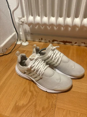 Vita Nike prestos - Snygga vita prestos från Nike med mesh-ovandel och tydlig swoosh-logga på tån. Skorna har vita snören, låg profil och en bekväm sula. Perfekta för en sportig och clean stil.  Tyvär små slitningar i hälarna på insidan 