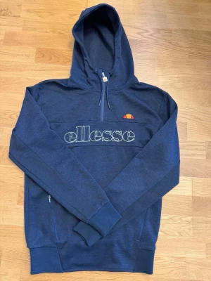 Mörkblå hoodie från Ellesse - Snygg mörkblå hoodie från Ellesse med halv dragkedja och stor logga broderad på bröstet. Klassisk huva och ribbade muddar vid ärmslut och nederkant. Praktiska fickor med dragkedja på sidorna. Perfekt för en avslappnad och sportig stil.