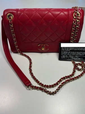Röd quiltad axelremsväska Chanel - Snygg röd handväska från Chanel i quiltat läder med guldfärgade detaljer och kedjerem. Klassisk CC-logga framtill och elegant design som ger lyxig känsla. Perfekt för dig som vill ha en ikonisk accessoar med tidlös stil. 0687