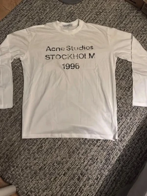 Vit långärmad t-shirt Acne Studios - Vit långärmad t-shirt från Acne Studios med tryck på bröstet där det står 'Acne Studios STOCKHOLM 1996'. Klassisk rund hals och relaxed fit. Perfekt för dig som gillar minimalistisk och stilren design.