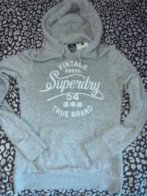 Grå Superdry hoodie med tryck - Snygg grå hoodie från Superdry med stort vitt tryck på bröstet där det står 'Vintage Goods Superdry 54 True Brand' och japanska tecken. Hoodien har huva med dragsko och en klassisk magficka. Perfekt för en avslappnad streetstyle-look.