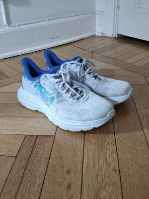 Ljusa Hoka Mach 4 sneakers med blå detaljer - Snygga ljusa sneakers från Hoka One Mach 4  med blå logga och blå insida. Skorna har en sportig design med mesh-ovandel för bra andningsförmåga och tjock, dämpad sula som ger skön komfort. Perfekta för dig som gillar streetstyle och sportig look. Storlek 42 i ny skick 