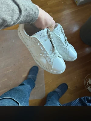 Vita sneakers med nitar - Säljer ett par vita sneakers med coola nitar längs sidorna och vita skosnören. Skorna har en klassisk låg profil och är tillverkade i slätt läder. Perfekta för dig som vill ha en stilren men edgy look. 1 nit har ramlat