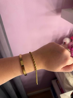 Guldigt Cordell armband  - Säljer två snygga armband i guldfärgad metall. Det ena är ett stelt armband med ingraverade symboler och det andra är ett tunnare tvinnat armband med enkel design. Perfekta accessoarer för att lyfta din stil.
