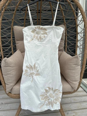 Klänning strl. 36 - Säljer en vit klänning med tunna axelband och broderade beige blommor framtill. På sidorna finns bälteshällor. Klänningen har en rak passform och slutar strax ovanför knäna.