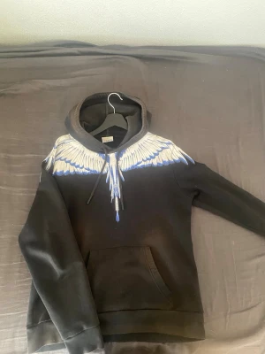 Marcelo burlon hoodie - Säljer en svart hoodie med stor huva och ett unikt vingmönster i vitt och blått över axlar och bröst, som fortsätter på ryggen. Hoodien har magficka framtill och dragsko i huvan. Perfekt för dig som vill sticka ut med en cool och kreativ design.