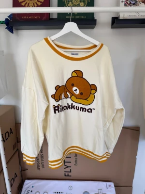 Gulvit Rilakkuma-tröja - Söt tröja i gulvitt med Rilakkuma-tryck framtill. Tröjan har gula ränder vid hals, ärmslut och nederkant samt en avslappnad passform och rund halsringning. Perfekt för dig som gillar kawaii-stil och mjuka plagg.