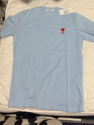 Ljusblå t-shirt med röd hjärtdetalj - Snygg ljusblå t-shirt med en liten röd hjärtdetalj och bokstaven A broderad på bröstet. Klassisk rund hals och korta ärmar. Perfekt för en clean och stilren look.