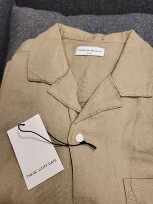 Beige linneskjorta These Glory Days - Snygg beige skjorta från These Glory Days i linne med korta ärmar och klassisk krage. Skjortan har knappar framtill och en bröstficka. Perfekt för varma dagar och enkel att styla till olika outfits.