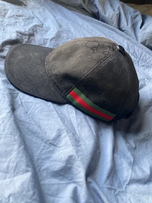 Svart keps med grön-röd rand - Snygg svart keps från Gucci med diskret GG-mönster i tyget och en klassisk grön-röd rand på sidan. Kepsen har böjd skärm och knapp på toppen. Perfekt accessoar för dig som gillar streetwear och vill ha något som sticker ut.