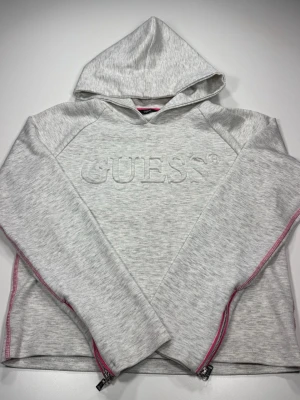 Ljusgrå hoodie från Guess med huva - Snygg ljusgrå hoodie från Guess med broderad logga framtill. Hoodien har dragkedjor i rosa längs sidorna och en klassisk huva. Tillverkad i mjukt sweatshirtmaterial och har långa ärmar. Perfekt för en avslappnad och trendig look.0172
