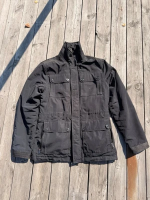 J Lindberg jacka  - Snygg svart jacka med hög krage och fyra stora fickor framtill, alla med knappstängning. Jackan har en rak passform och justerbara detaljer baktill. Perfekt för blåsiga dagar och enkel att matcha med olika outfits.