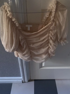 Beige volangblus med puffärm - Supersöt beige blus med volanger och puffärmar. Toppen har omlottkänsla, rynkade detaljer och är tillverkad i ett tunt, transparent material med små prickar. Perfekt för dig som vill ha en romantisk och trendig look.