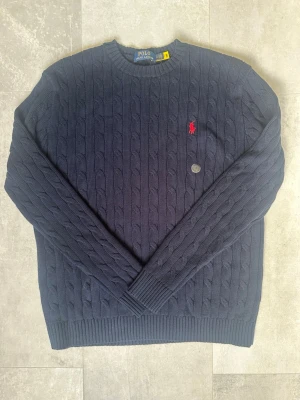Mörkblå stickad tröja Polo Ralph Lauren - Kabelstickad mörkblå tröja från Polo Ralph Lauren med klassisk röd broderad logga på bröstet. Perfekt för dig som gillar stilren och tidlös design. Den är bara använd någon gång. Passar även du som har storlek M 
