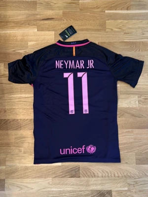 Barça Neymar Jr matchtröja lila - Snygg FC Barcelona matchtröja i mörklila med rosa detaljer, Neymar Jr och nummer 11 på ryggen. Framsidan har Qatar Airways-logga och klubbmärke, samt Champions League-patchar på ärmarna. Perfekt för dig som älskar fotboll och vill sticka ut på planen.