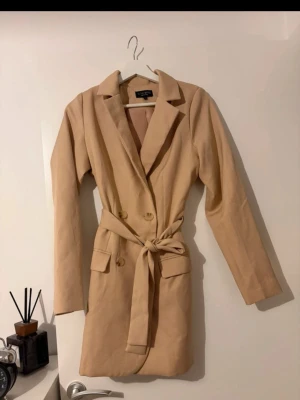 Beige kavajklänning - Stilren beige kavajklänning med knappar framtill och matchande bälte i midjan. Klänningen har lång ärm, krage och fickdetaljer, vilket ger en elegant och trendig look. Perfekt för dig som gillar klassiska plagg med modern twist.