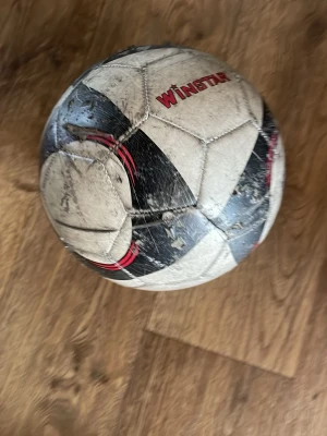 Vit och svart fotboll från Winstar - Säljer en klassisk fotboll från Winstar i vitt och svart med röda detaljer. Bollen har traditionellt mönster med sexkanter och är perfekt för träning eller spontanspel med vänner. Materialet är slitstarkt syntetläder.