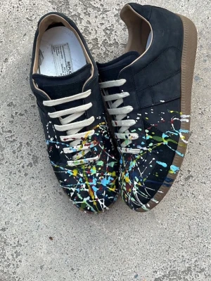 Maison Margiela GATS - Unika svarta sneakers med färgstänk i blått, gult, grönt och vitt. Skorna har beige snörning och brun sula. Materialet ser ut att vara mocka eller nubuck. Perfekta för dig som vill sticka ut med din stil.