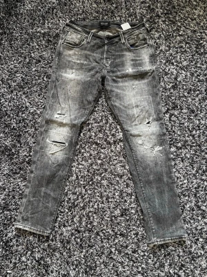 Grå slitna skinny jeans - Säljer ett par grå skinny jeans med slitningar och distressed detaljer på både fram- och baksida. Jeansen har fem fickor, dragkedjegylf och en cool tvättad look. Perfekta för dig som gillar en avslappnad och trendig stil.