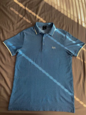 Blå pikétröja från Hugo Boss - Snygg blå pikétröja från Hugo Boss med vit logga broderad på bröstet. Tröjan har klassisk krage med vit kant, knappar framtill och korta ärmar med vita detaljer vid ärmsluten. Perfekt till en avslappnad men stilren look.