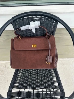 Mörkbrun handväska  - Snygg mörkbrun handväska i vegansk mocka med detaljer i slätt läder. Väskan har ett klassiskt top handle, guldfärgat lås och dekorativ rem med hängande detalj. Perfekt storlek för det viktigaste och ger en elegant touch till din outfit.