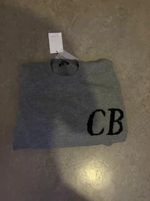 Grå stickad tröja med CB-detalj - Snygg grå stickad tröja med rund hals och ribbade muddar. På framsidan finns en svart CB-detalj som ger en cool touch. Perfekt för dig som gillar enkel men trendig stil.