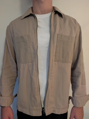 Beige overshirt/skjortjacka, stl S (passar M) - Beige overshirt i bomullstwill, två bröstfickor och dragkedja hela vägen upp. Zara, stl S men avslappnad passform så den funkar fint som M också. Går att style öppen över en t-shirt eller stängd som en lätt jacka. Bra skick. Modell: 182cm | 83kg.