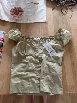 Ljusgrön blus med puffärmar - Supersöt ljusgrön blus med puffiga kortärmar och knappar framtill. Offshoulder-modell med rynkade detaljer vid halsringningen. Perfekt för dig som vill ha en feminin och trendig topp till sommaren.