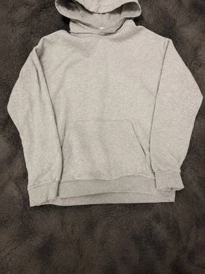 Grå hoodie från H&M - En enkel och stilren grå hoodie från H&M med huva och magficka. Tillverkad i mjukt bomullsmaterial och har ribbade muddar vid ärmslut och nederkant. Perfekt för en avslappnad och bekväm look.