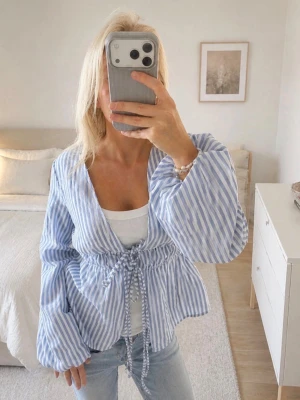 Blårandig blus med knytband - Snygg blårandig blus med v-ringning och knytband i midjan. Blusen har långa, vida ärmar och är tillverkad i ett lätt och luftigt material. Perfekt att styla med jeans för en fräsch look.