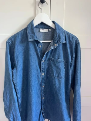 Blå jeansskjorta från B.M.D - Klassisk blå jeansskjorta från B.M.D med lång ärm, bröstficka och knappar framtill. Skjortan har en avslappnad passform och är tillverkad i ett mjukt jeanstyg som passar perfekt till både jeans och chinos. Storlek 170