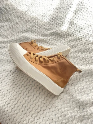 Beige höga sneakers med RL-logga - Snygga beige sneakers med högt skaft och RL-logga på sidan. Skorna har vita, rejäla gummisulor och tåhätta samt matchande beige snörning. Ovandelen är i canvas och mockaliknande material vid hälen. Perfekta för en trendig och avslappnad stil.                                                    