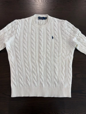 Vit kabelstickad tröja - Kabelstickad vit tröja med rund halsringning och klassisk marinblå logga broderad på bröstet. Tröjan har ribbade muddar vid ärmslut och nederkant. Perfekt för en stilren och tidlös look. Sitter som S/m