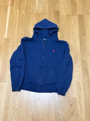 Mörkblå hoodie från Polo Ralph Lauren - Mörkblå hoodie med dragkedja från Polo Ralph Lauren. Klassisk design med huva, känguruficka och röd broderad logga på bröstet. Tillverkad i mjukt bomullsmaterial och har ribbade muddar vid ärmar och nederkant. Perfekt för en avslappnad och stilren look.