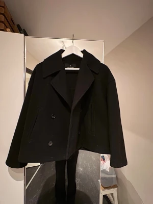 Kort svart ullkappa med dubbelknäppning - Stilren svart kappa i ull med dubbelknäppning och bred krage. Modellen är kort och har raka ärmar samt två fickor framtill. Perfekt för dig som vill ha en klassisk och trendig look till hösten eller vintern.