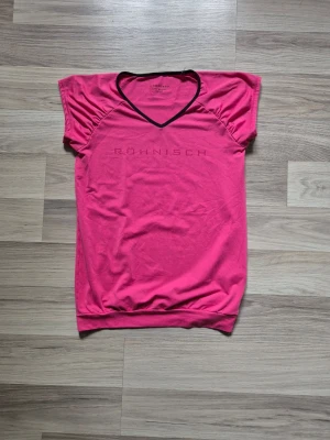 Rosa träningstopp från Röhnisch med v-ringning - Snygg rosa träningstopp från Röhnisch med svart v-ringad kant och korta ärmar. Toppen har ett sportigt snitt och är tillverkad i ett mjukt material som passar perfekt till träning eller en aktiv vardag.