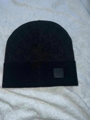 Svart ribbad mössa med patch - Snygg svart mössa med ribbad kant och en liten fyrkantig patch framtill. Klassisk och enkel design som passar till det mesta. Mössan är stickad och har en uppvikt kant för extra stilpoäng.